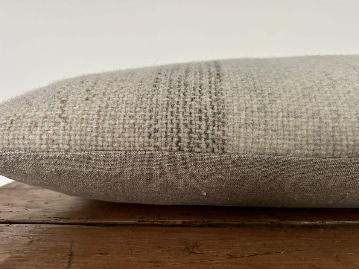 Side view of the woven wool cushion showing the grey stripe detail and the natural linen backing, on a wooden surface