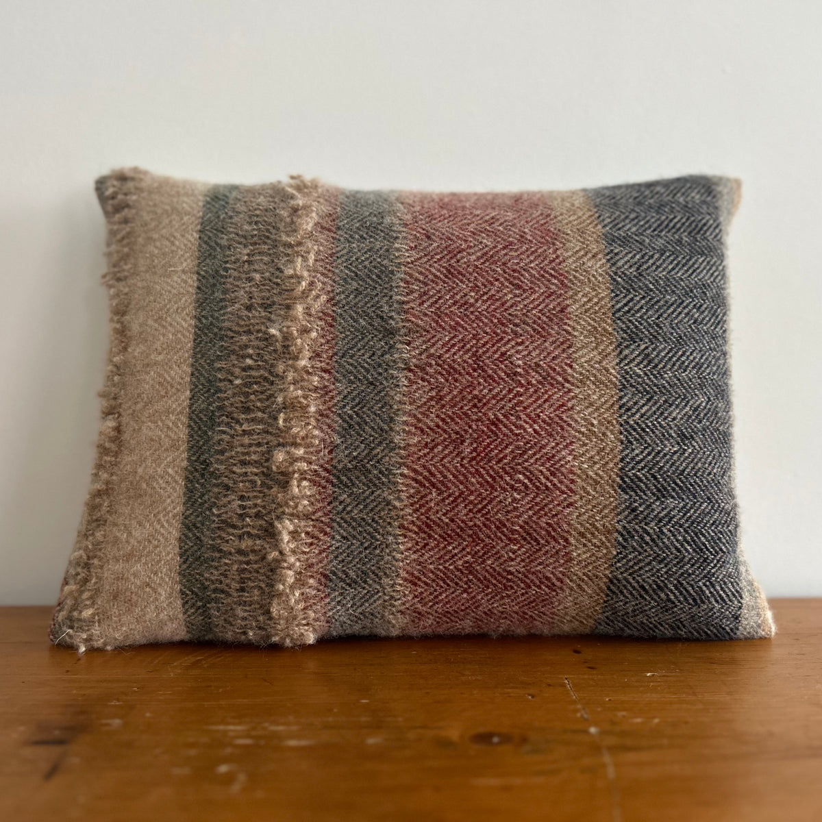 Rectangular wool cushion woven in vertical stripes of oatmeal, grey-green, brick red and navy herringbone, with a raised band of looped yarn a third of the way across