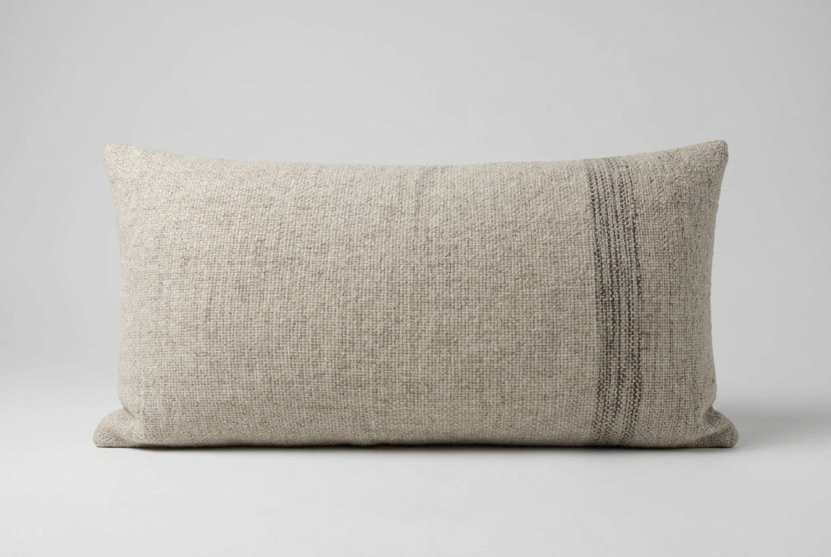 Rectangular oatmeal-grey woven wool cushion with a narrow band of dark grey stripes near one end, against a plain grey background